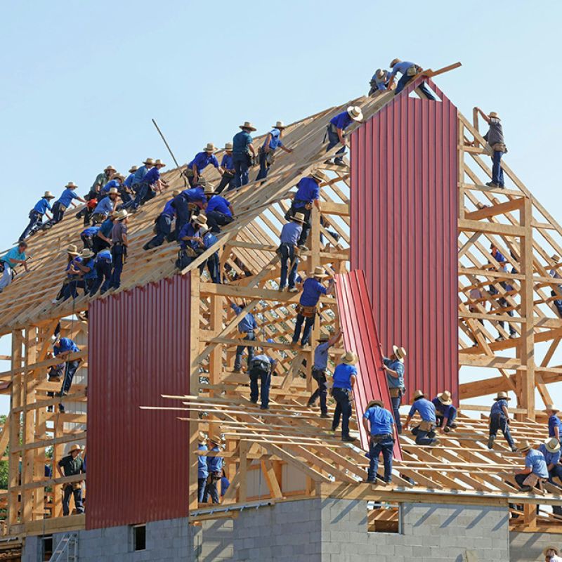 Viele Menschen bauen gemeinsam eine Scheune – Symbol für Teamarbeit, Struktur und gelingende Transformation.